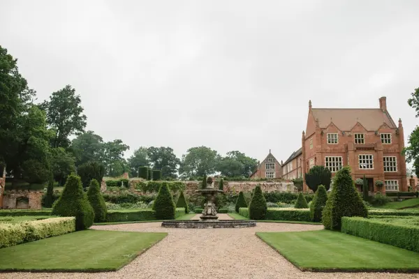 Lush, manicured gardens surround a grand, historic brick manor, featuring elegant hedges and a central fountain for outdoor ceremonies.