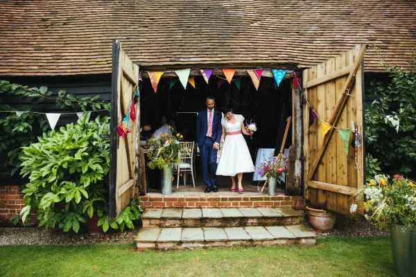 Charming rustic barn entrance adorned with colorful bunting, leading to a cozy celebration space with greenery and guests.