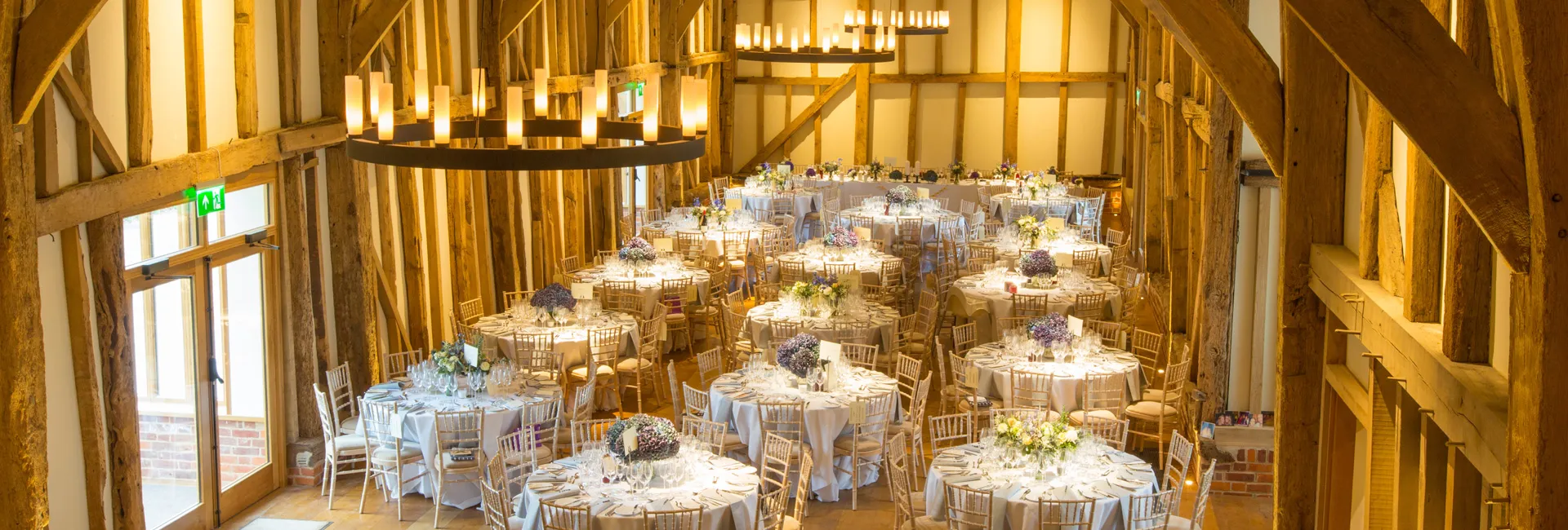 Bright and airy rustic barn featuring elegantly set round tables adorned with floral centerpieces and warm wooden beams above.