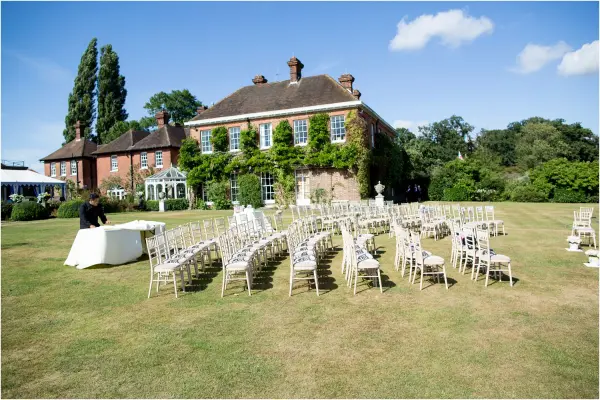 Charming outdoor ceremony space featuring white chairs arranged on manicured lawn, with a picturesque brick venue and lush greenery backdrop.