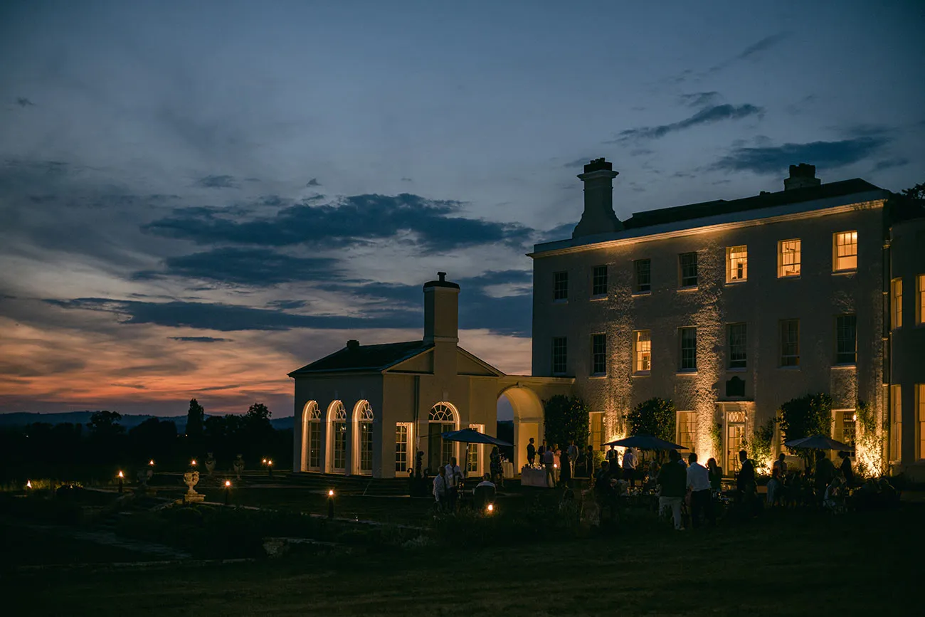 Elegant estate venue at twilight, featuring beautifully lit walls, outdoor seating areas, and a breathtaking sunset backdrop perfect for weddings.