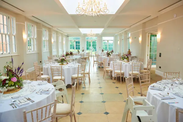 Elegant reception space featuring round tables with floral centerpieces, natural light from large windows, and a stunning chandelier overhead.