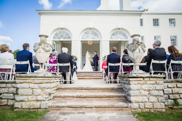 Charming outdoor wedding ceremony set on stone steps, flanked by elegant urns, with guests seated under a bright blue sky.