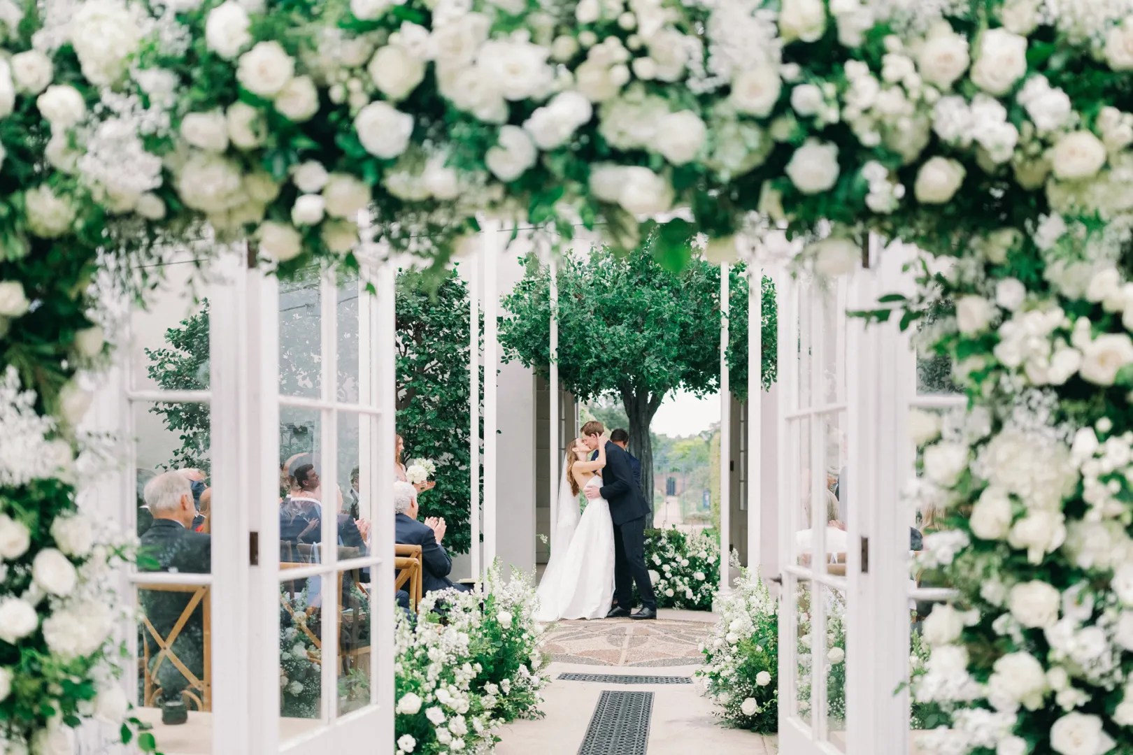 An elegant ceremony under a floral archway, featuring lush white roses and greenery, with a couple exchanging vows nearby.