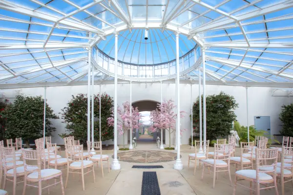 Bright indoor ceremony space featuring elegant rows of chairs under a glass ceiling, framed by lush greenery and delicate pink blossoms.