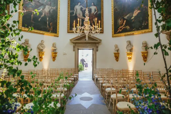 Elegant indoor ceremony space featuring golden chandeliers, classical artwork, and rows of wooden chairs surrounded by greenery.