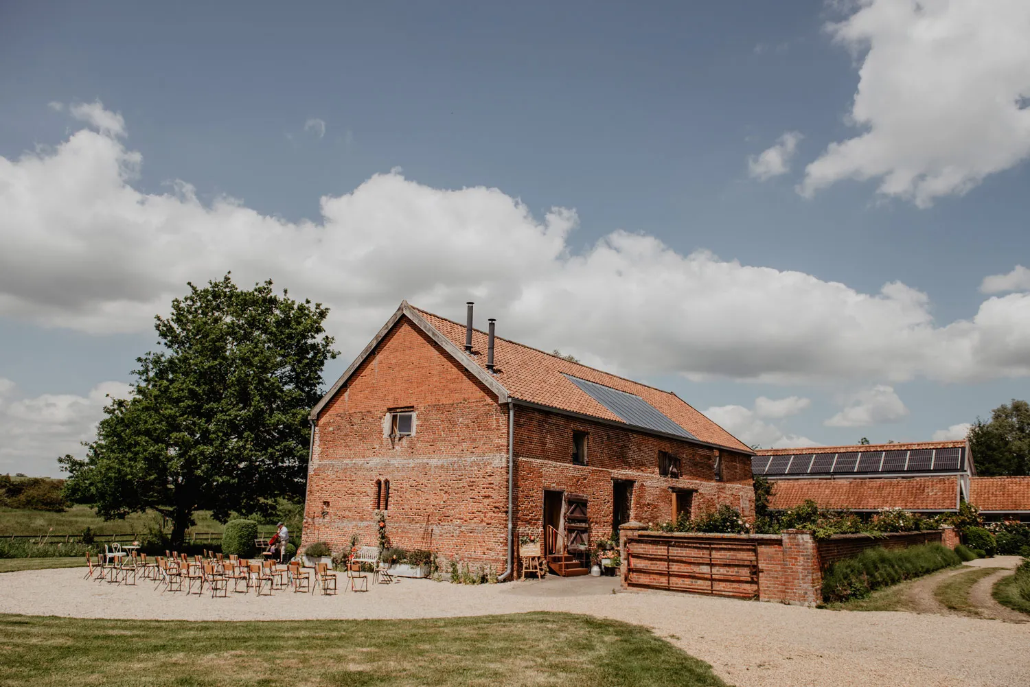 Charming rustic barn set amidst green fields, featuring a gravel path and a ceremony space lined with chairs under a vibrant sky.