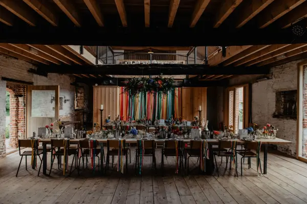 Stylish reception space with a long, decorated table, vibrant floral arrangements, and colorful ribbon accents under warm wooden beams.