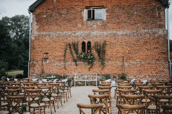Charming outdoor ceremony space featuring wooden chairs arranged in front of a rustic brick wall adorned with greenery and floral accents.