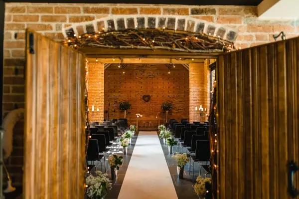 Rustic wooden doors open to reveal a warm, intimate ceremony space with brick walls, elegant seating, and floral decorations lining the aisle.