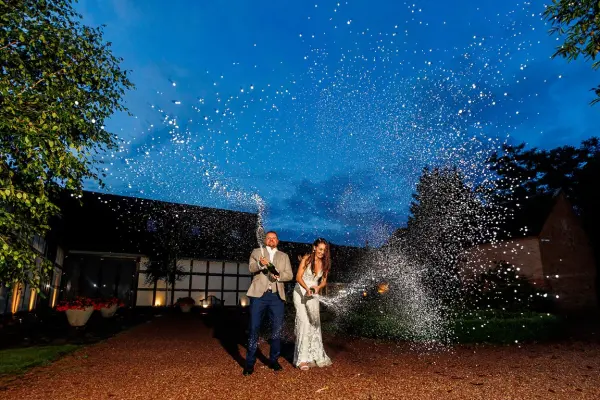 Couple celebrating with sparklers outside a charming venue, illuminated by evening light, surrounded by lush greenery and rustic architecture.