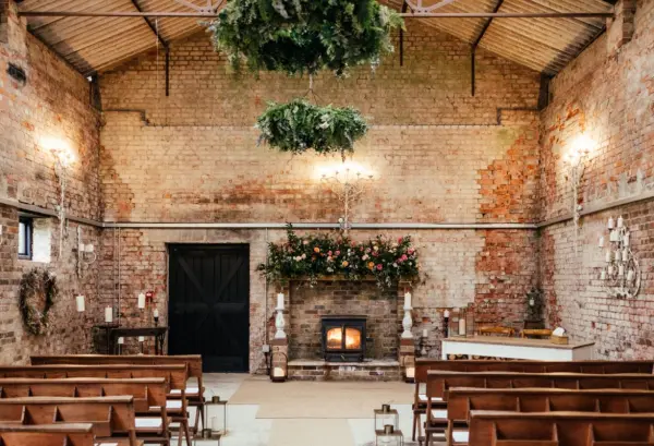 Charming rustic ceremony space with exposed brick walls, wooden seating, and elegant floral accents above a cozy fireplace.