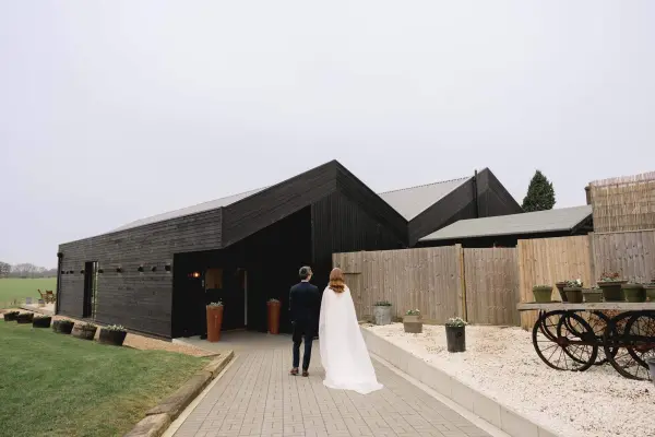 A couple walks towards a modern black barn venue, surrounded by greenery and rustic decor, perfect for a charming wedding celebration.
