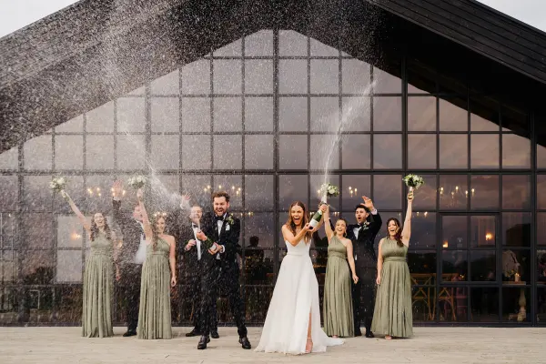Bridal party celebrating outside a modern venue with large glass windows, holding bouquets and spraying confetti in the air.