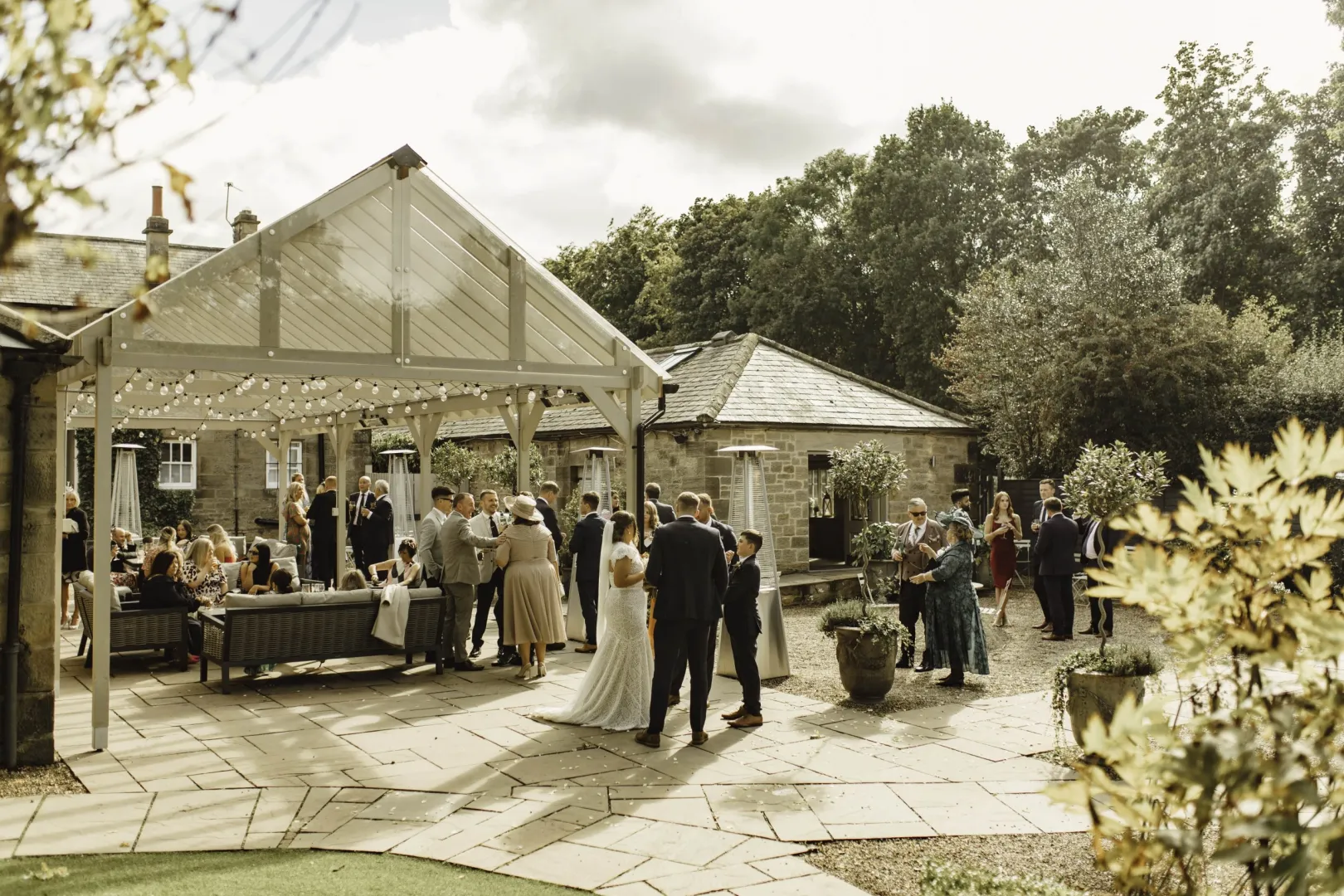 Charming outdoor reception area with rustic decor, string lights, and a lively gathering of wedding guests on a sunny day.