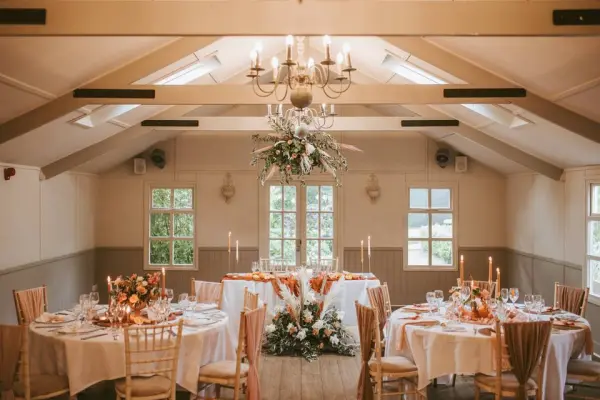 Charming reception space featuring elegantly set tables, rustic chandelier, and floral decorations under a bright, airy ambiance.