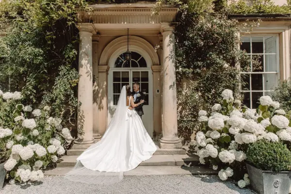 Elegant couple at a grand entrance surrounded by blooming hydrangeas and lush greenery, perfect for a romantic wedding backdrop.