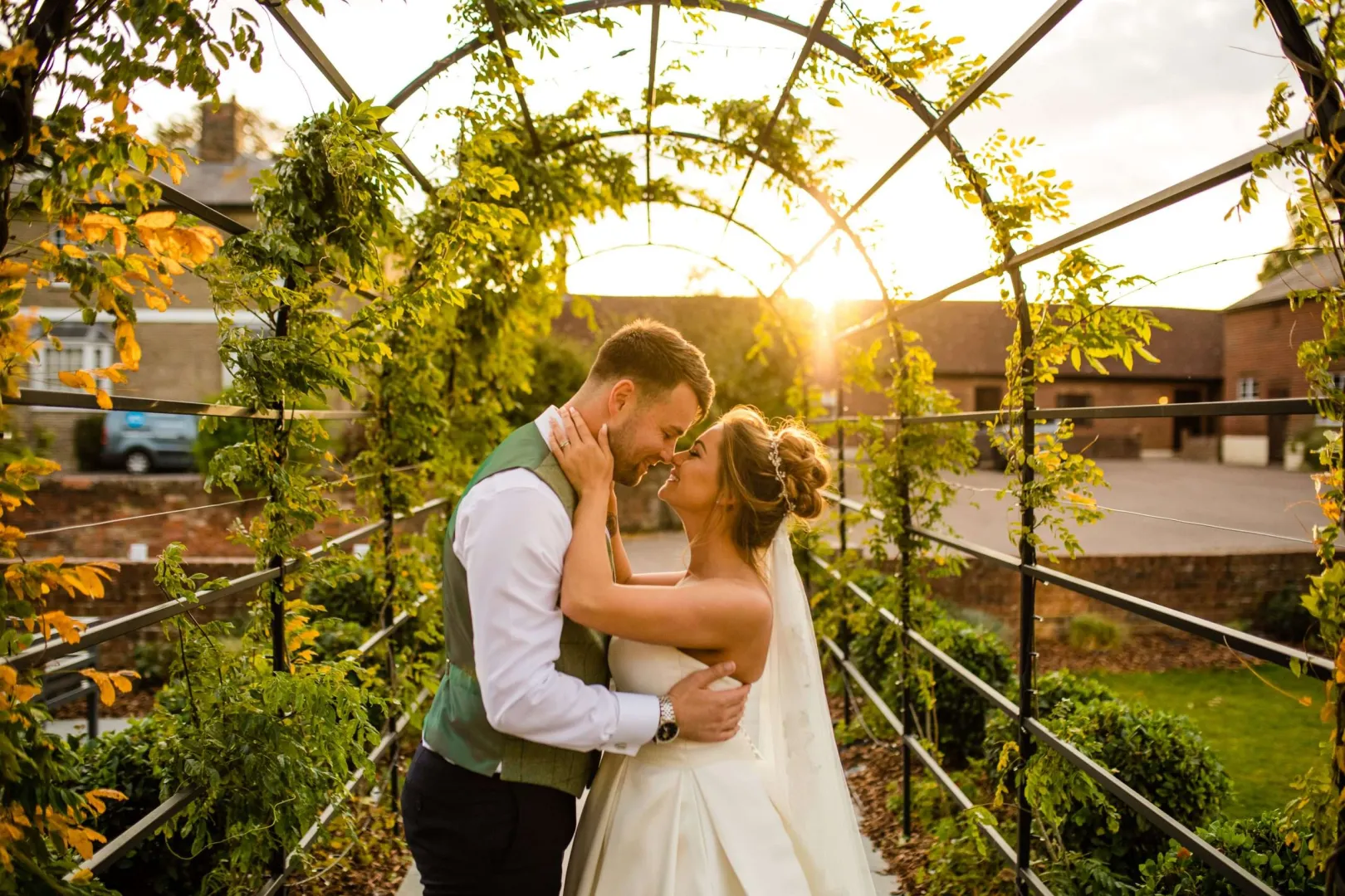 Couple embracing in a charming garden archway adorned with greenery, backlit by a warm, golden sunset. Perfect for romantic outdoor ceremonies.