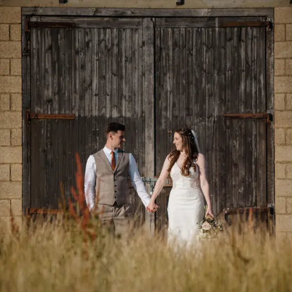 A couple stands hand-in-hand in front of rustic wooden doors, surrounded by golden grasses, capturing a romantic outdoor wedding moment.