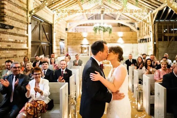 Rustic barn wedding venue with wooden beams and charming decor, featuring a couple sharing their first kiss while guests applaud.