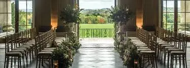Elegant indoor ceremony space adorned with floral arrangements, lined with chairs, showcasing expansive views of lush greenery outside.