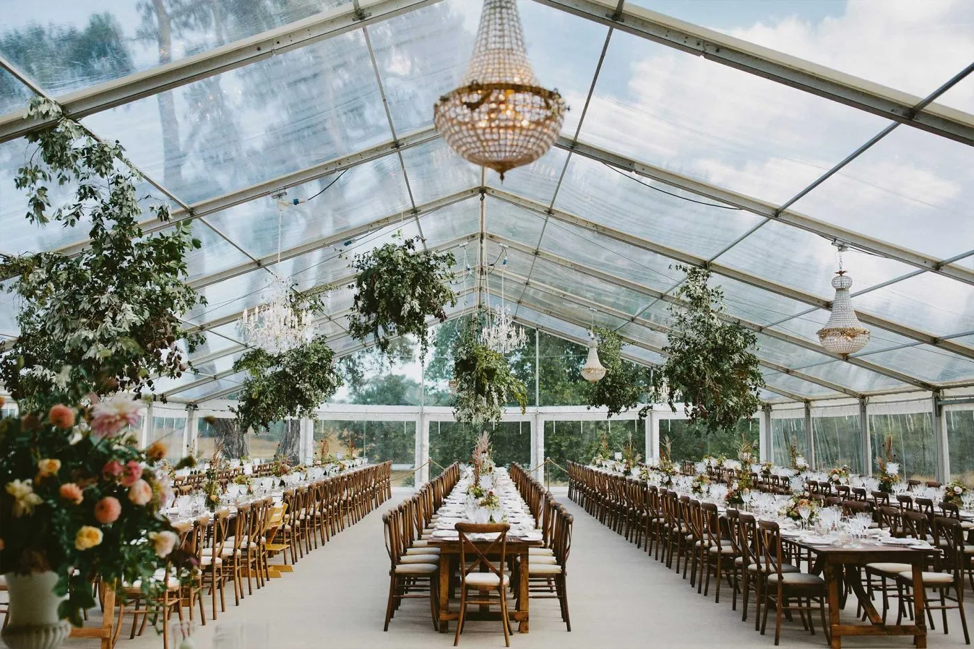 Stylish glass tent venue with elegant chandeliers, long reception tables, and lush greenery, perfect for a romantic wedding celebration.