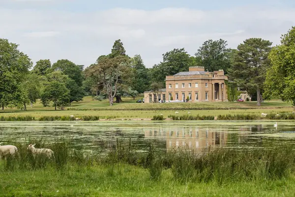 Charming venue featuring a grand estate near a serene lake, surrounded by lush greenery and grazing sheep, ideal for outdoor weddings.