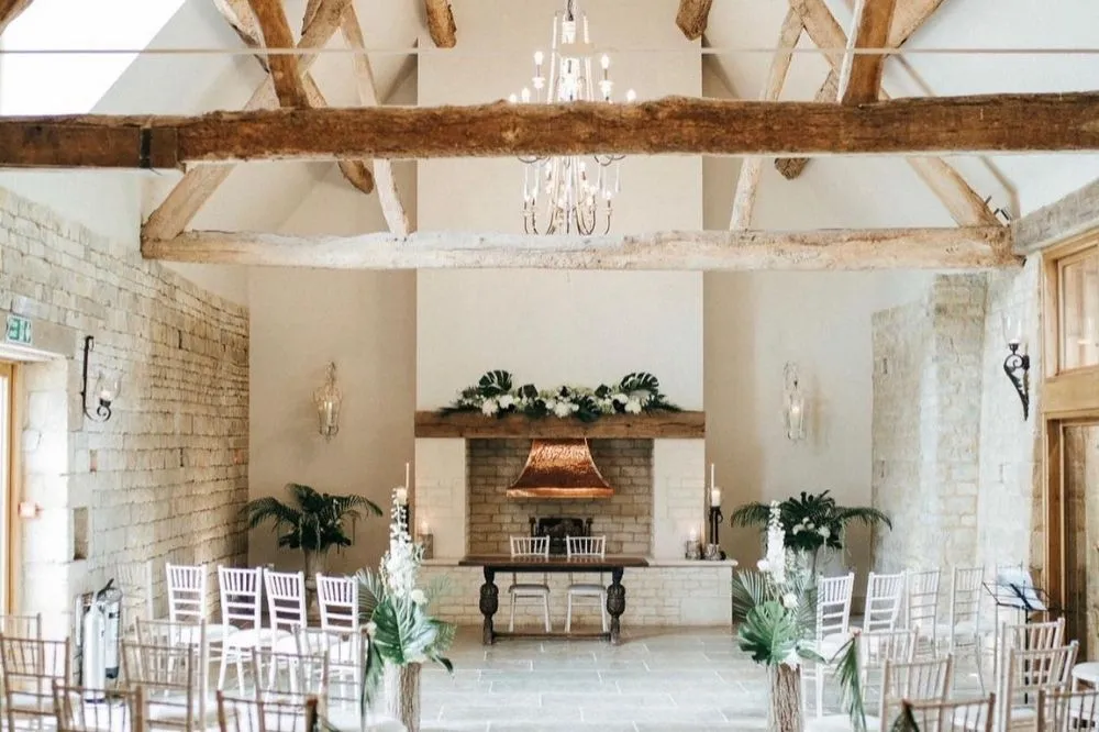 Spacious indoor wedding venue with rustic beams, elegant chandelier, a charming fireplace, and rows of chairs ready for guests.