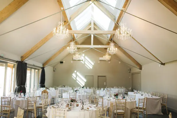 Spacious reception hall features elegant chandeliers, wooden beams, and round tables set for a wedding celebration.