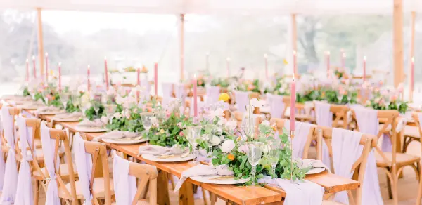 Long wooden tables adorned with lush greenery, elegant glassware, and soft floral centerpieces, set under a bright event tent.