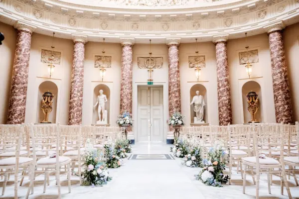 Elegant ceremony space featuring classical statues, pink marble columns, and rows of wooden chairs adorned with floral arrangements.