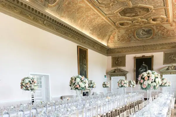 Elegant ballroom with ornate ceiling, long tables set for a wedding, and stunning floral centerpieces in tall vases.