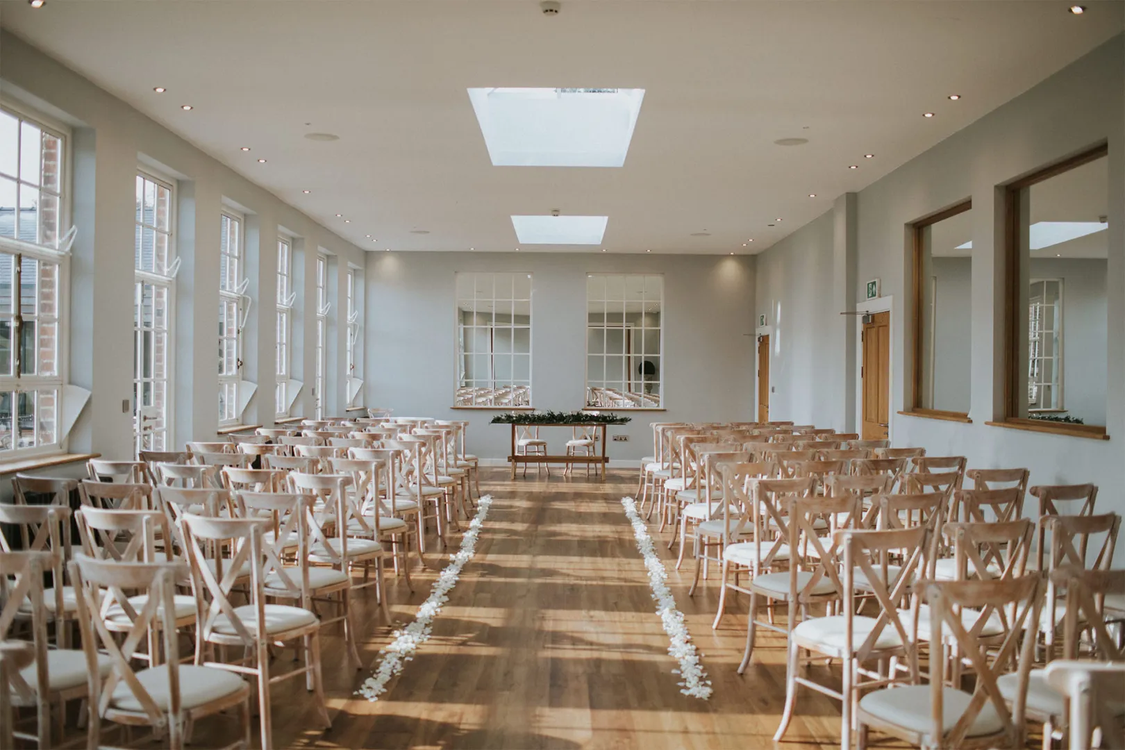 Bright, airy ceremony space featuring elegant wooden chairs arranged in neat rows, large windows, and a central focal mirror.