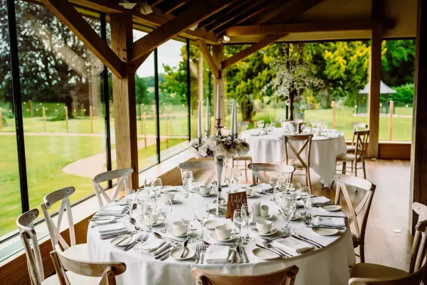 Bright, rustic dining area with elegant tables set for a wedding, featuring large windows, greenery outside, and wooden beams.