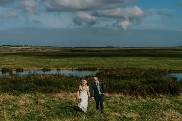 Couple walking hand in hand through lush green fields, with serene water in the background and a clear blue sky above. Ideal for outdoor weddings.