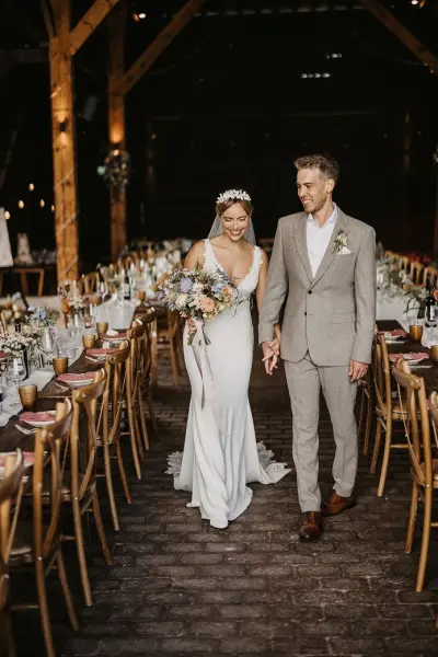 A bride and groom walk hand in hand through a rustic barn, featuring long reception tables set with elegant floral arrangements.