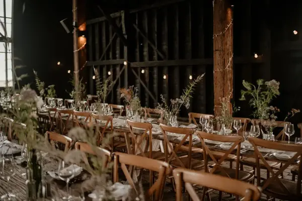 A rustic barn interior featuring long wooden tables set with elegant dinnerware and delicate floral arrangements, illuminated by soft lighting.