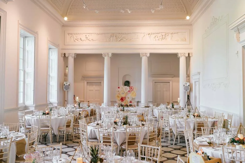 Elegant ballroom featuring round tables adorned with white linens, sparkling centerpieces, and intricate architectural details. Bright natural light filters through tall windows.