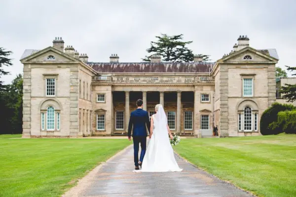 Newlyweds stroll hand-in-hand down a path toward a grand stone mansion, surrounded by lush green lawns and towering trees.