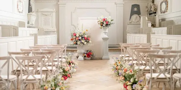 Charming indoor ceremony space featuring white decor, wooden crossback chairs, and vibrant floral arrangements lining the aisle.