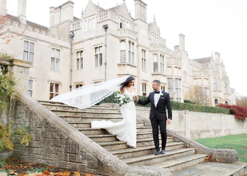A couple ascends grand stone steps, with an elegant estate backdrop, adorned in wedding attire, showcasing a romantic outdoor setting.