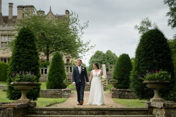 A bride and groom stroll hand-in-hand along a landscaped path, framed by lush greenery and elegant topiary in a serene garden setting.