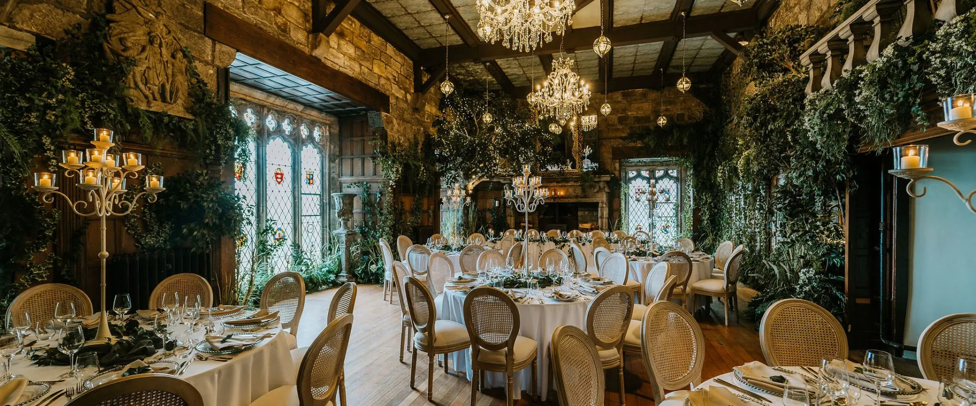 Elegant reception space featuring rustic stone walls, chandeliers, and lush greenery, with round tables set for a romantic celebration.