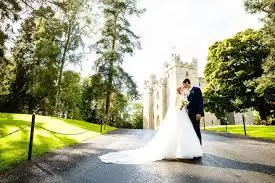 A couple embraces on a tree-lined path leading to a romantic castle, framed by lush greenery and soft sunlight, perfect for weddings.