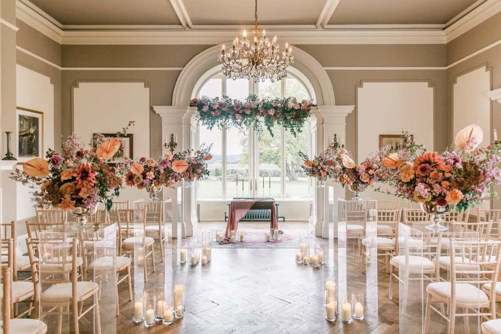 Elegant ceremony space featuring soft floral arrangements, candlelight, and a grand chandelier, all framed by large windows.