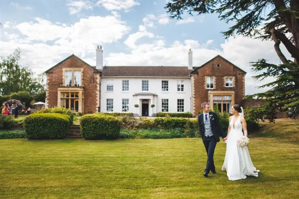 A couple strolls hand in hand across a lush lawn, highlighting a charming historic mansion with elegant architecture and picturesque gardens.