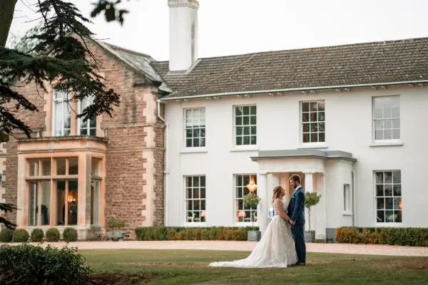 A couple stands hand-in-hand in front of a charming white mansion, featuring elegant architecture and manicured gardens, perfect for weddings.