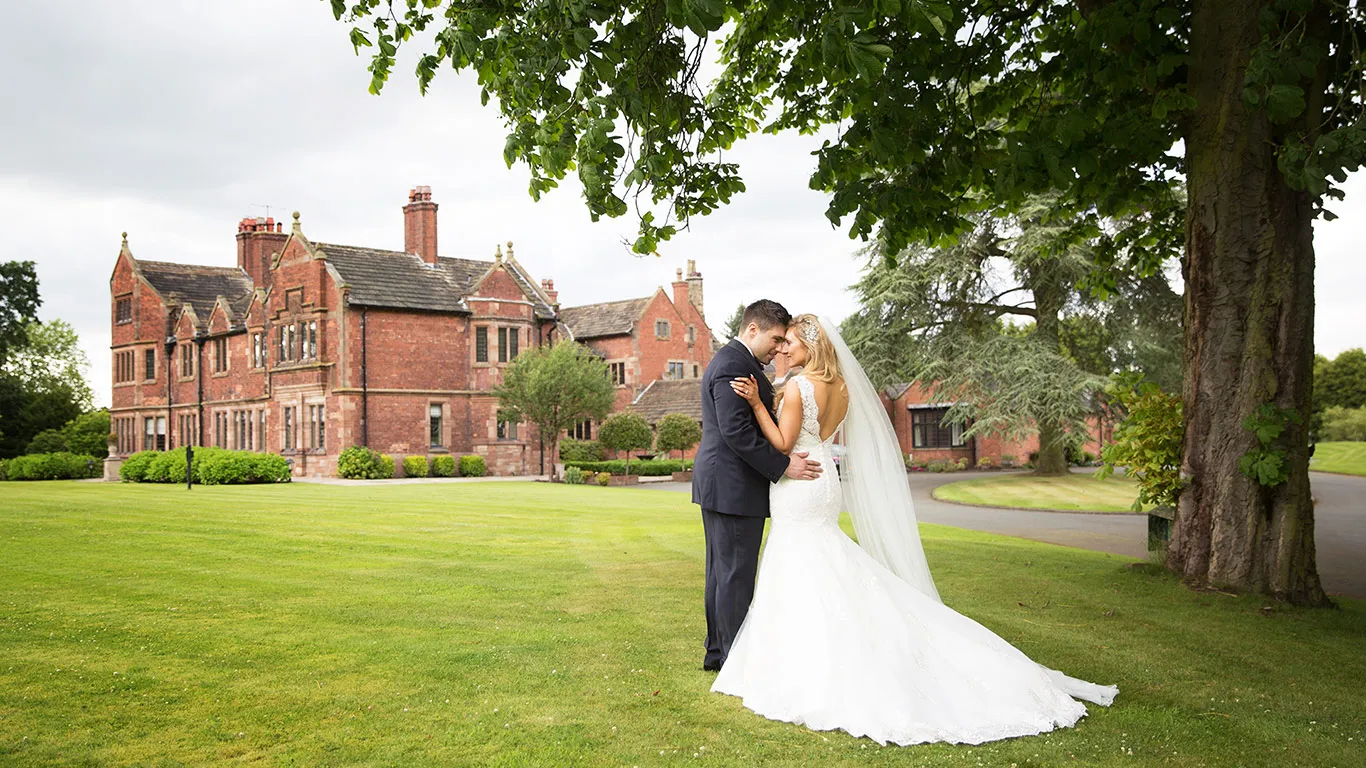 A bride and groom embrace on manicured lawns, framed by a grand red brick mansion and lush greenery, ideal for elegant outdoor ceremonies.