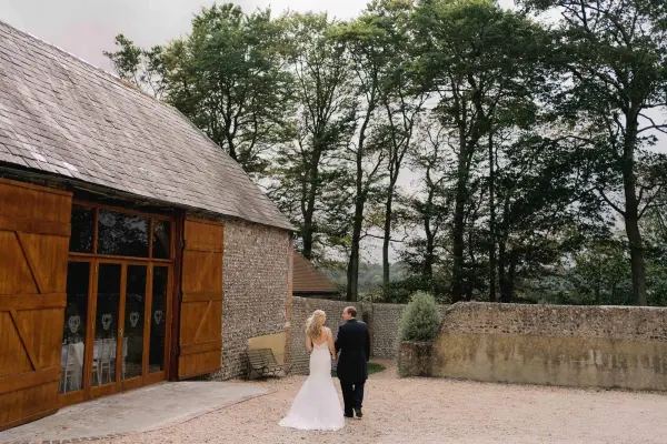 Couple walking hand-in-hand near rustic barn venue, surrounded by lush trees and a serene, open courtyard. Perfect for an intimate wedding.