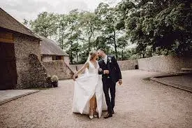 A bride and groom stroll hand-in-hand along a gravel path, surrounded by lush greenery and rustic stone buildings, perfect for a charming wedding.